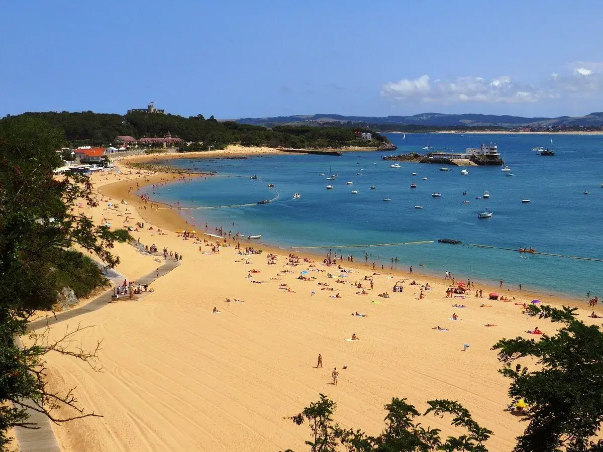 La **playa de los peligros** se llena de gente disfrutando del sol y el mar azul, con barcos anclados y edificios en la costa.