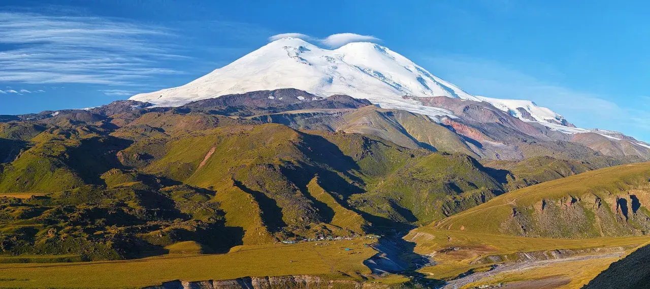 Elbrus szczyt w chmurach, otoczony zielonymi zboczami i obozem u podnóża.