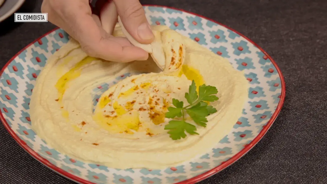 Un trozo de pan pita se sumerge en un cremoso hummus, adornado con aceite de oliva y perejil. Descubre qué es el tahini y cómo prepararlo.