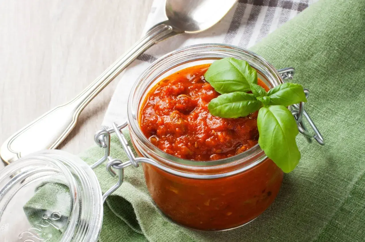 Tarro de cristal con salsa napolitana casera, decorado con hojas de albahaca fresca. Cuchara plateada y servilleta verde al lado.