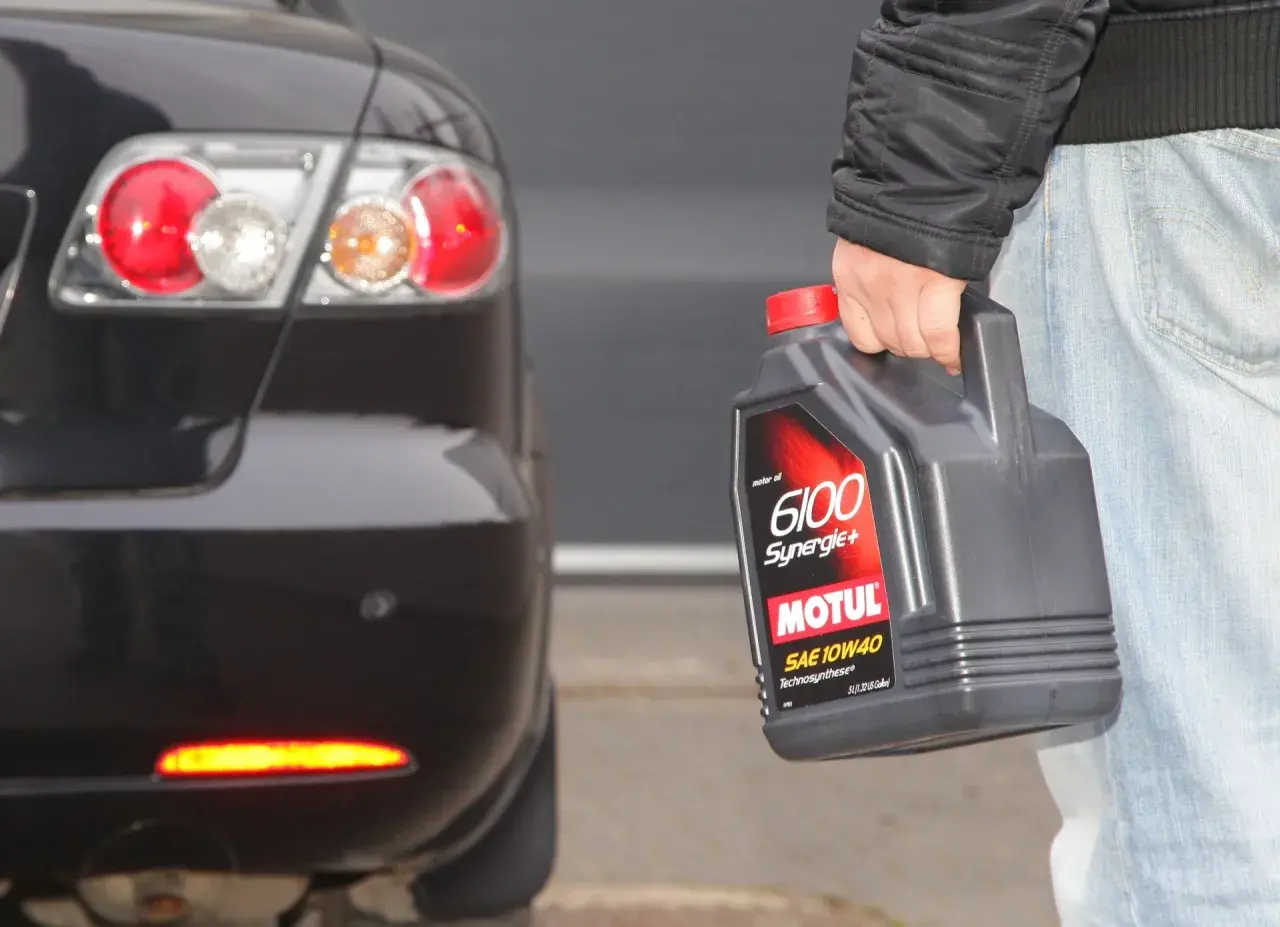 Osoba trzyma olej Motul 6100 Synergie+ SAE 10W40 obok tylnej części czarnego samochodu. Idealny wybór, by dobrze dobrać olej.