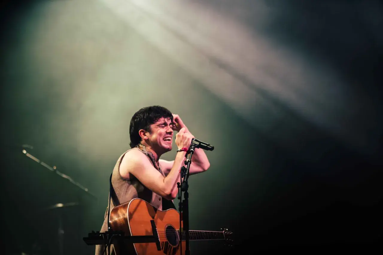 Declan McKenna passionately sings into a microphone, guitar in hand, bathed in stage lights. A glimpse of his setlist is visible.
