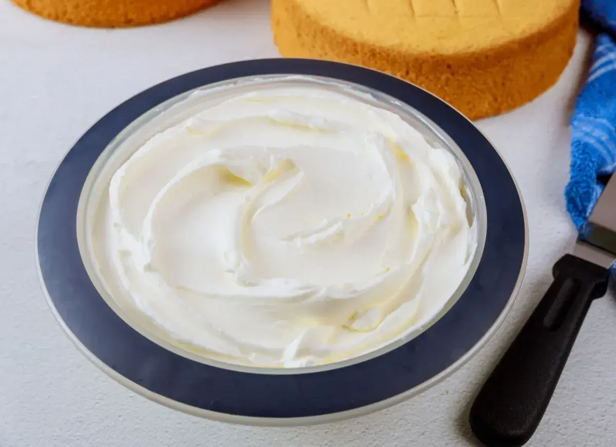 Puszysta bita śmietana z mascarpone w misce, gotowa do dekorowania tortu. Obok leżą biszkopty i szpatułka.