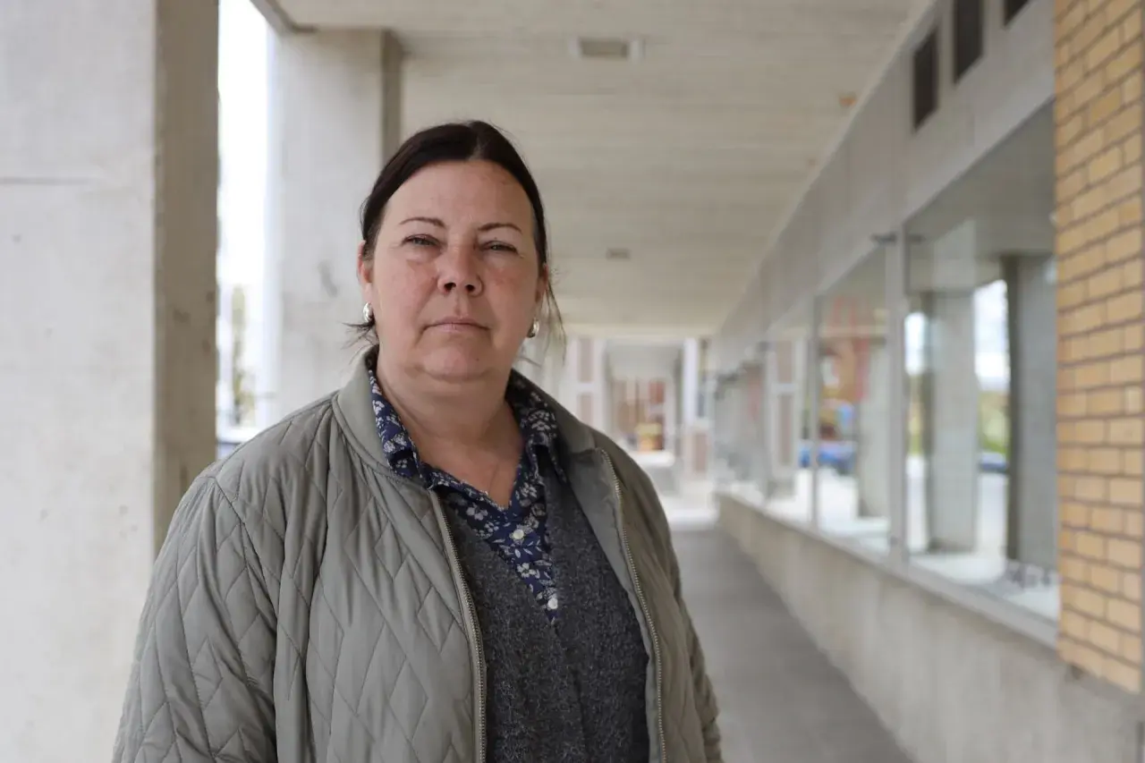 Jessica de Geer står utanför en byggnad med tegel och betong. Hon bär en grå jacka över en mönstrad skjorta och en stickad väst.