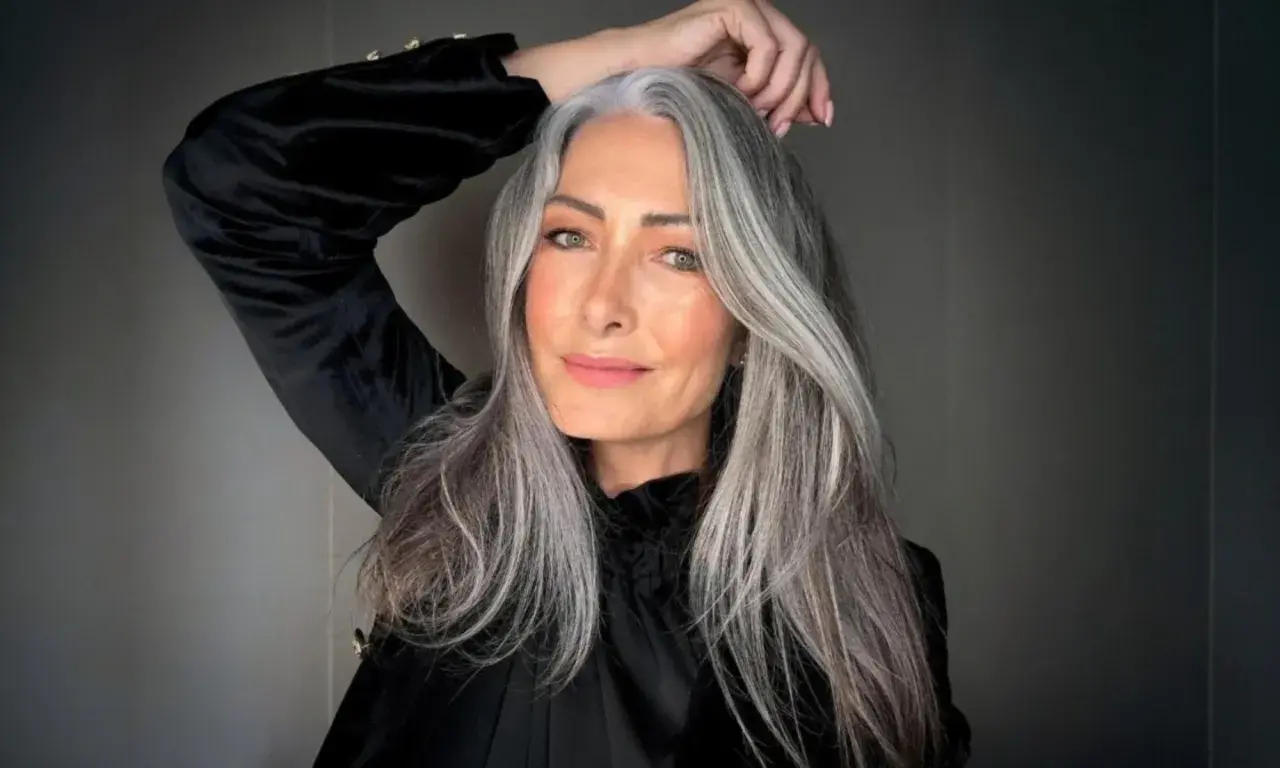 Mujer con cabello largo y ondulado, mostrando una hermosa integración de canas. Su mano acaricia su cabeza, resaltando la textura y el color natural.