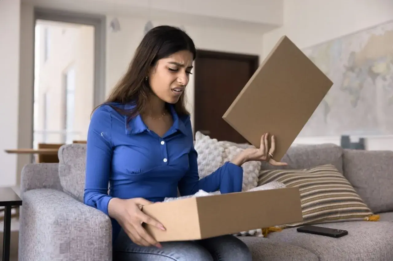 Femme déçue ouvrant un colis, découvrant un produit non conforme à la commande. Elle exprime son mécontentement.