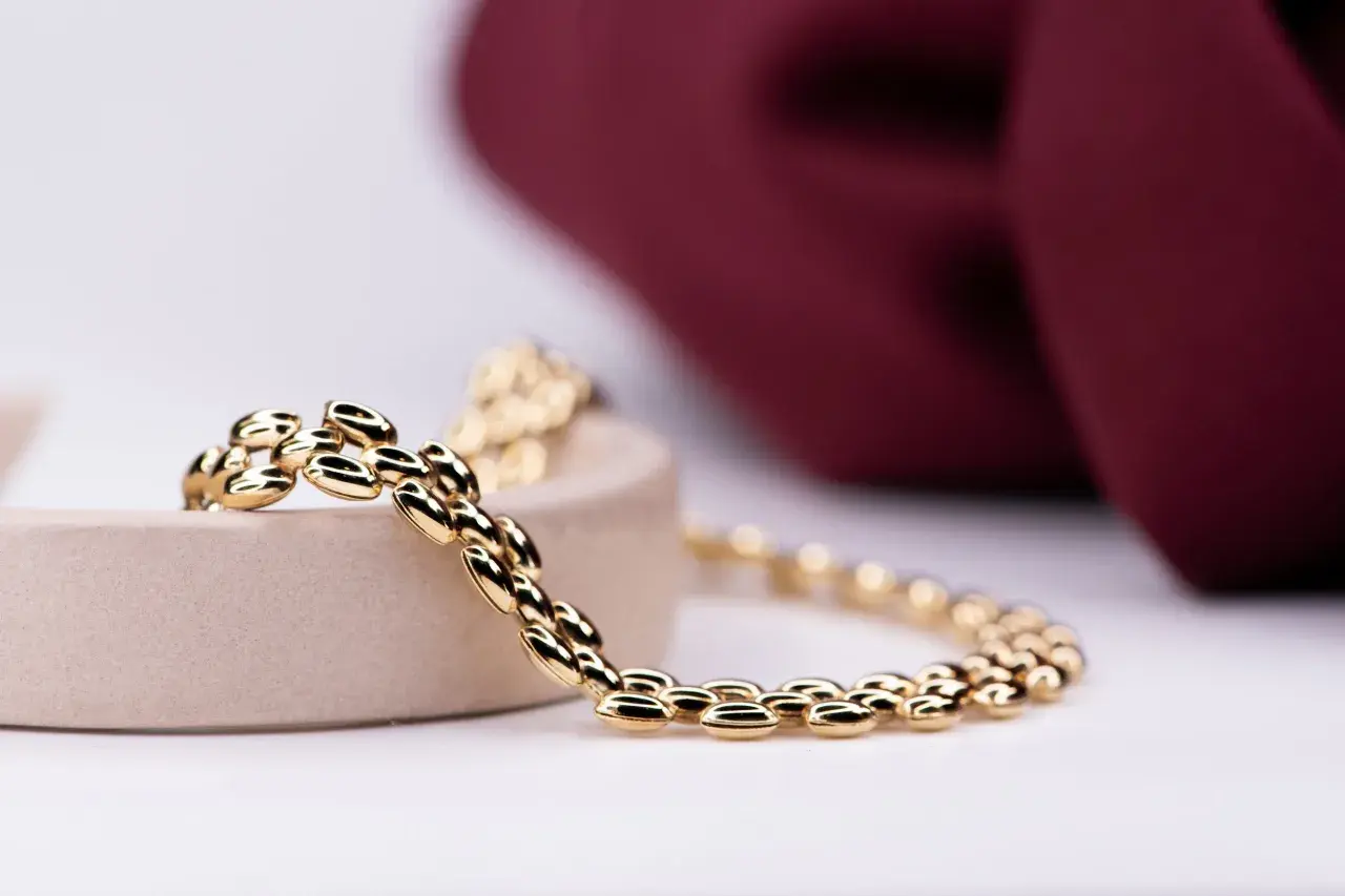 Pulsera de eslabones ovalados en oro, brillando sobre un soporte beige, con un fondo de tela burdeos.