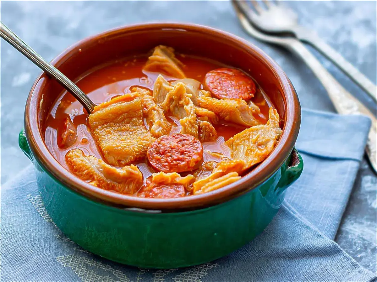 Un tazón de barro con callos a la madrileña, trozos de chorizo y un caldo rojizo. Una cuchara metálica está dentro.