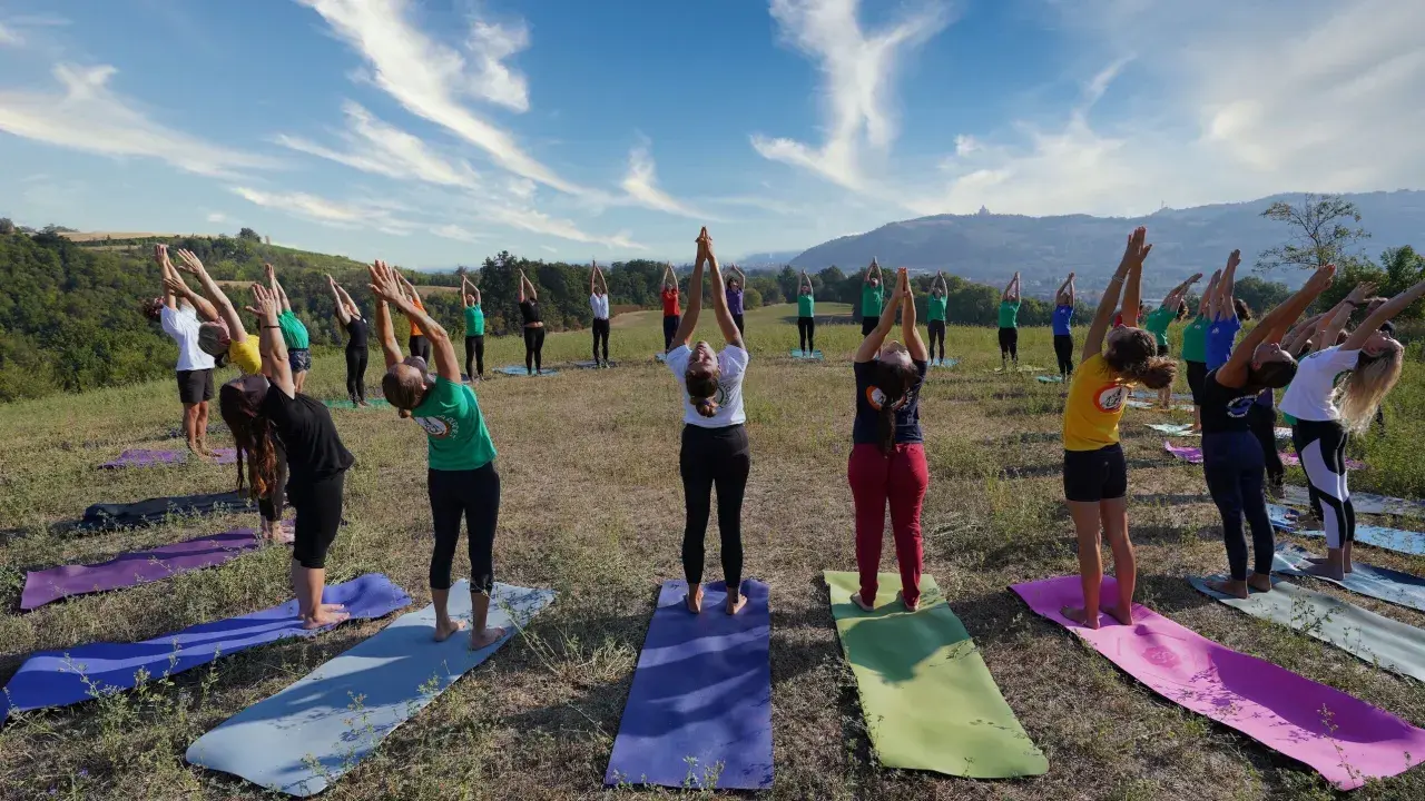 Gruppo di persone in posa urdhva hastasana su tappetini da yoga in un prato verde, con cielo azzurro e nuvole bianche.