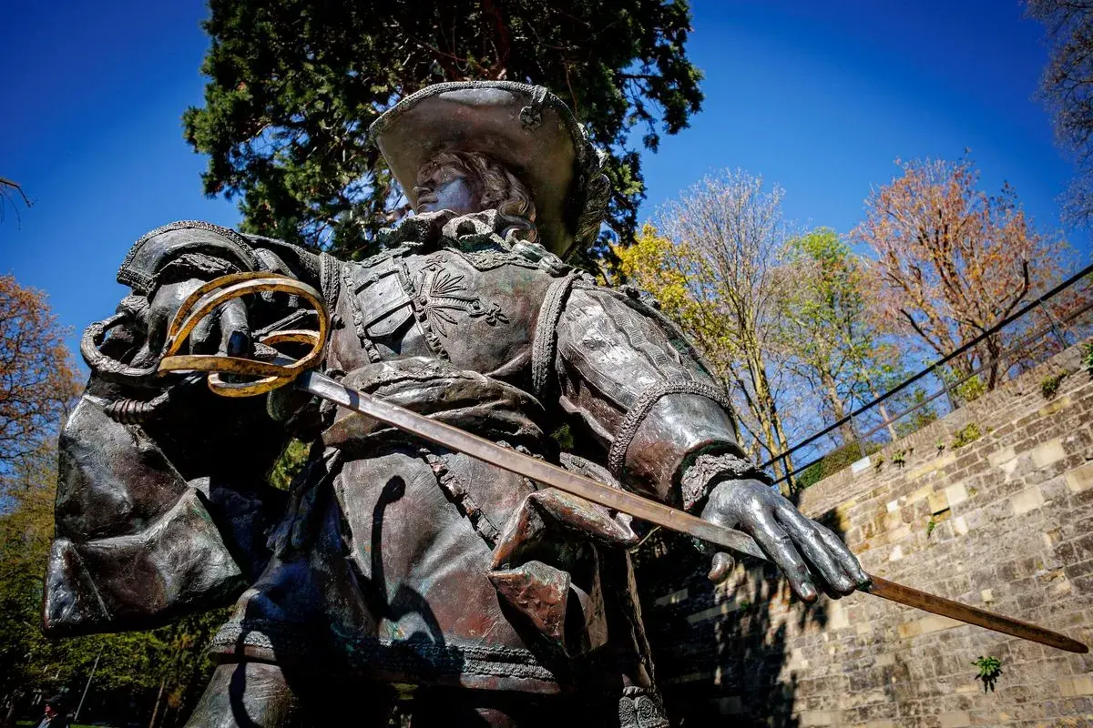 Bronzestatue eines Musketiers bei Dumas, mit Schwert und Hut, vor blauem Himmel und Bäumen.
