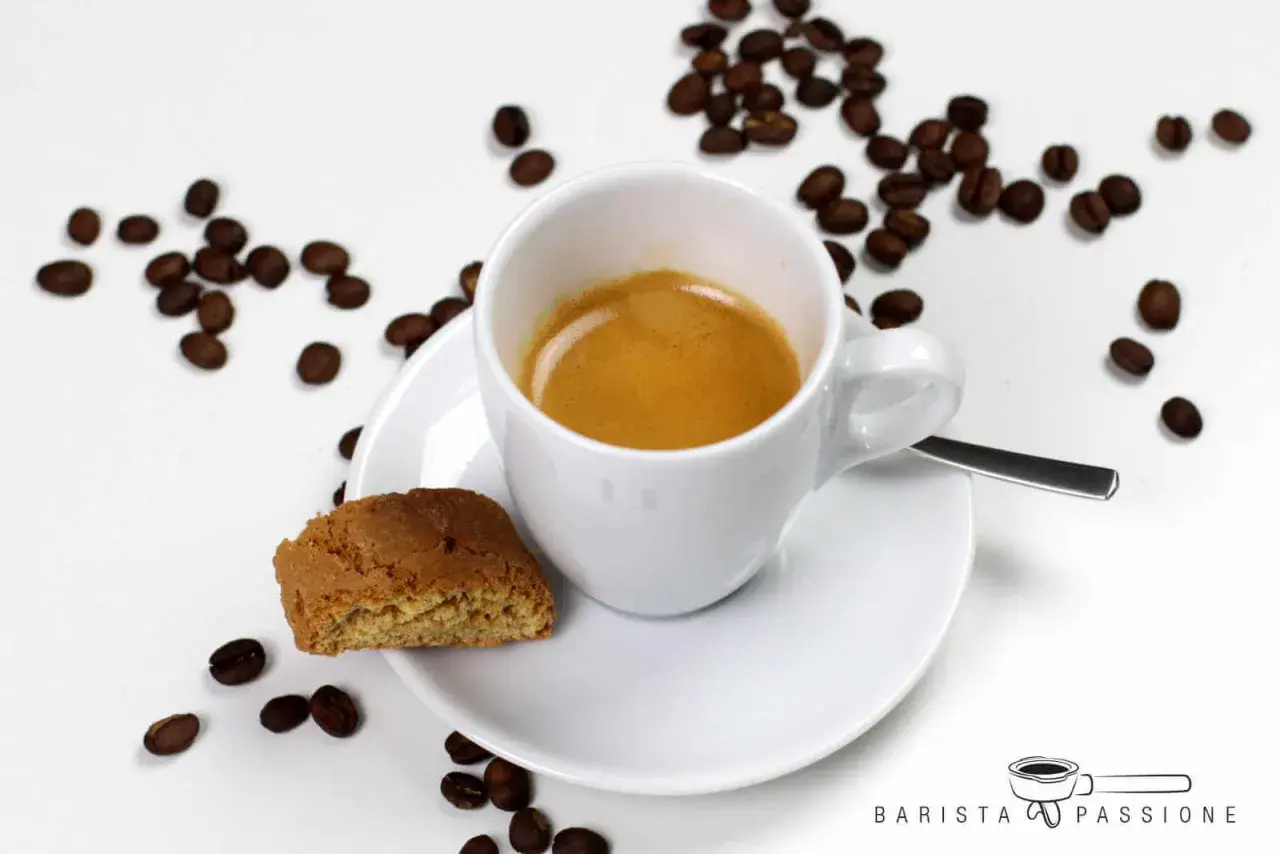 Ein weißer Becher mit Kaffee, daneben ein Cantuccini und Kaffeebohnen. Das ist ein Kaffee Lungo, ein längerer Espresso.