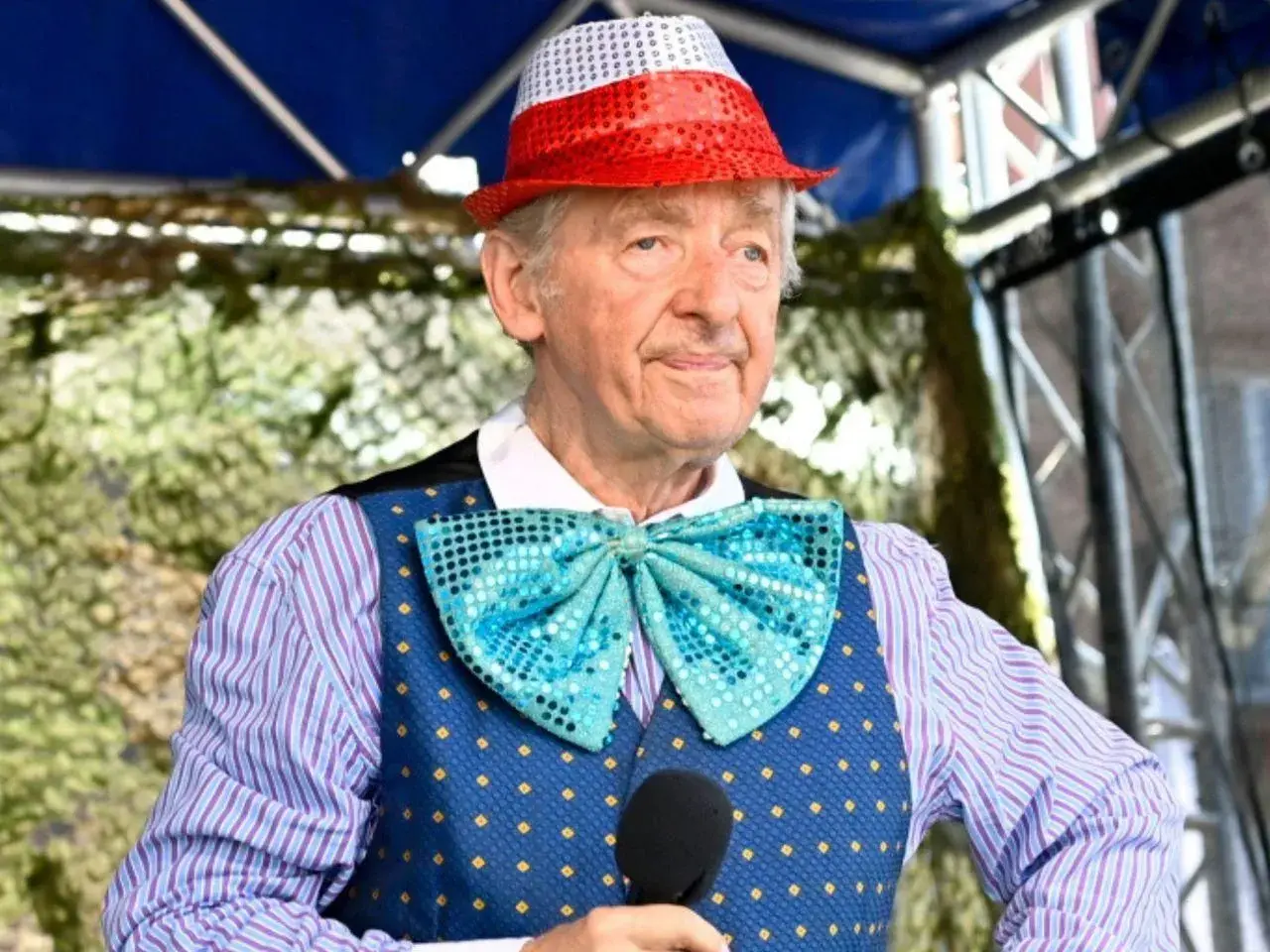 Uśmiechnięty Andrzej Rosiewicz w cekinowym kapeluszu i muszce, trzymający mikrofon. Wiek artysty to 80 lat.
