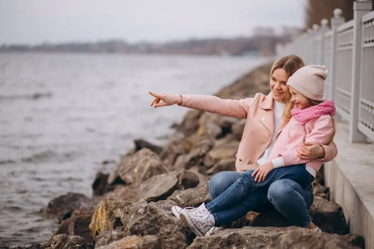 Mama z córką siedzą na kamieniach nad jeziorem. Mama wskazuje coś palcem, a dziewczynka się śmieje. To świetne gąski atrakcje dla dzieci!