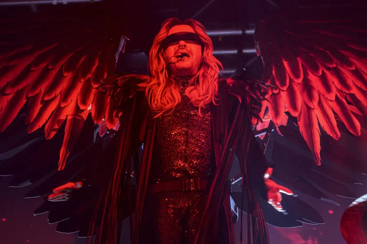Bill Kaulitz z Tokio Hotel na koncercie w anielskich skrzydłach i błyszczącym stroju, w czerwonym świetle.