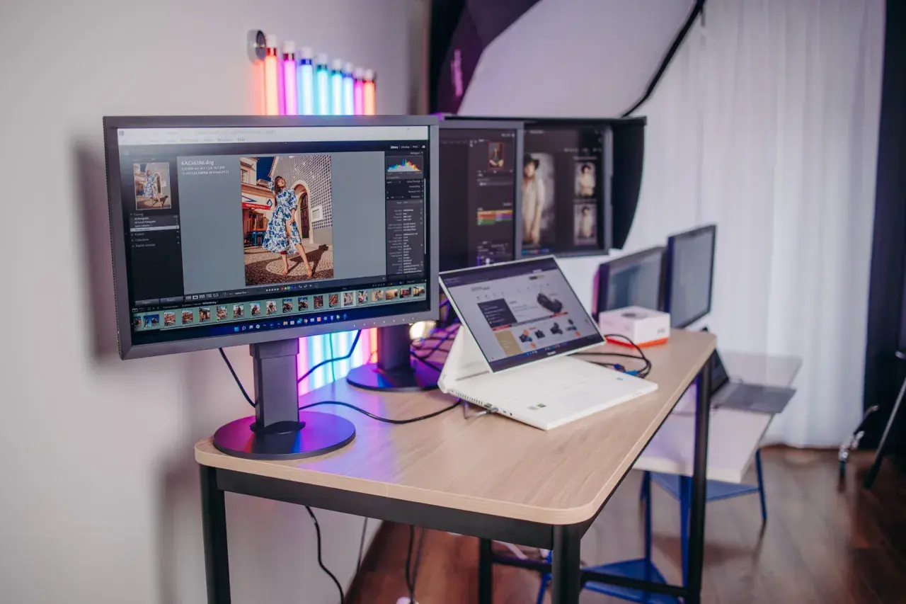 Profesjonalny monitor do obróbki zdjęć, z programem Lightroom, pokazuje zdjęcie kobiety w sukience. Obok laptop i inne monitory.
