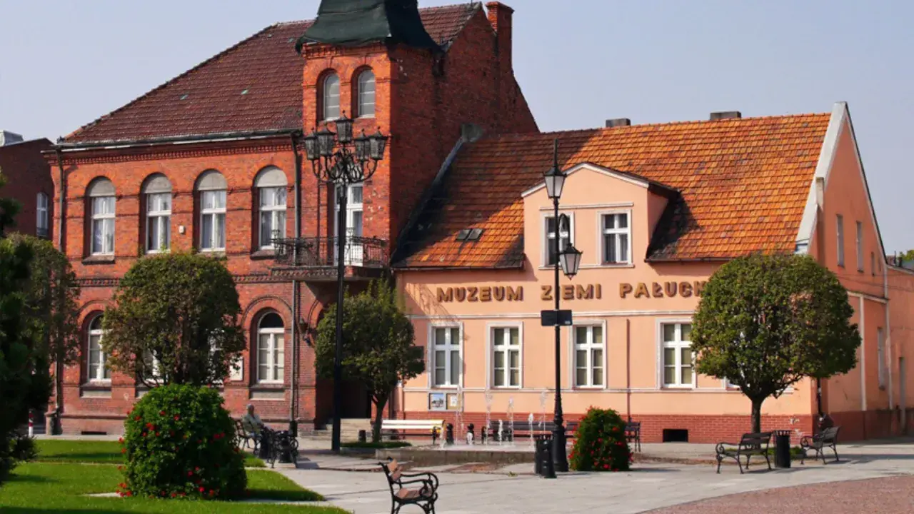 Muzeum Ziemi Pałuckiej, z ceglaną fasadą i pomarańczowym budynkiem, otoczone zielenią i fontanną.