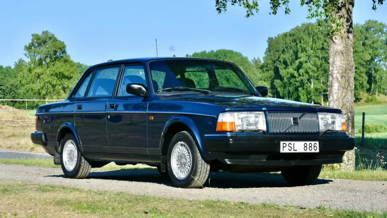 Mörkblå Volvo 240 från 1993 parkerad på en grusväg med grönskande träd och fält i bakgrunden.