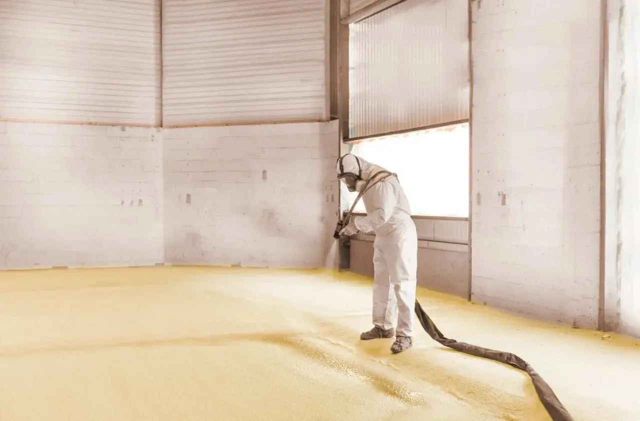 Un ouvrier en combinaison projette de la mousse polyuréthane sur le sol d'un entrepôt pour une isolation efficace.