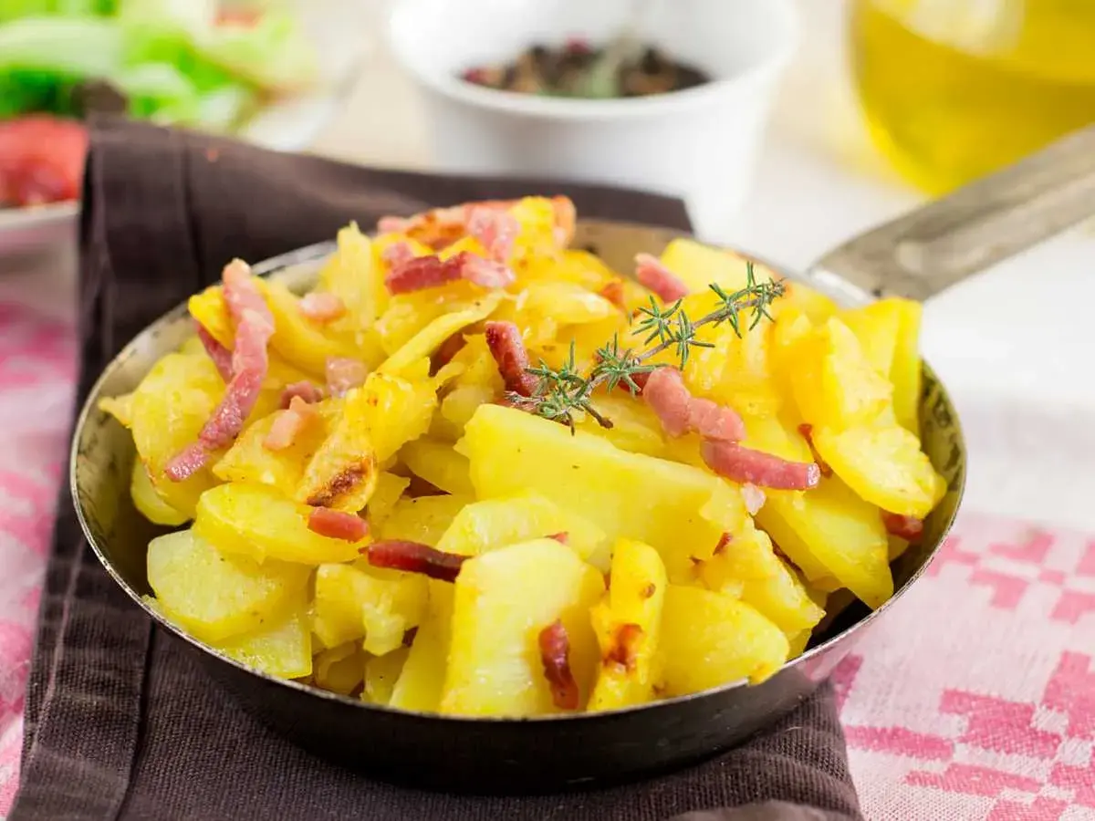 Patatas al microondas con trocitos de bacon crujiente y romero fresco, servidas en una sartén metálica.