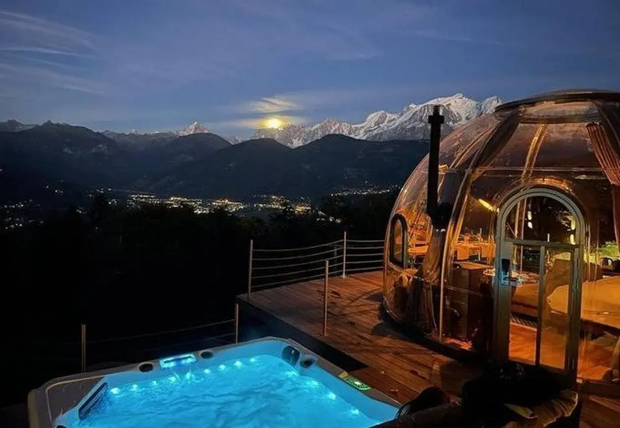 Un jacuzzi illuminé et une bulle transparente, un logement insolite montagne offrant une vue spectaculaire sur les sommets enneigés et la vallée scintillante.