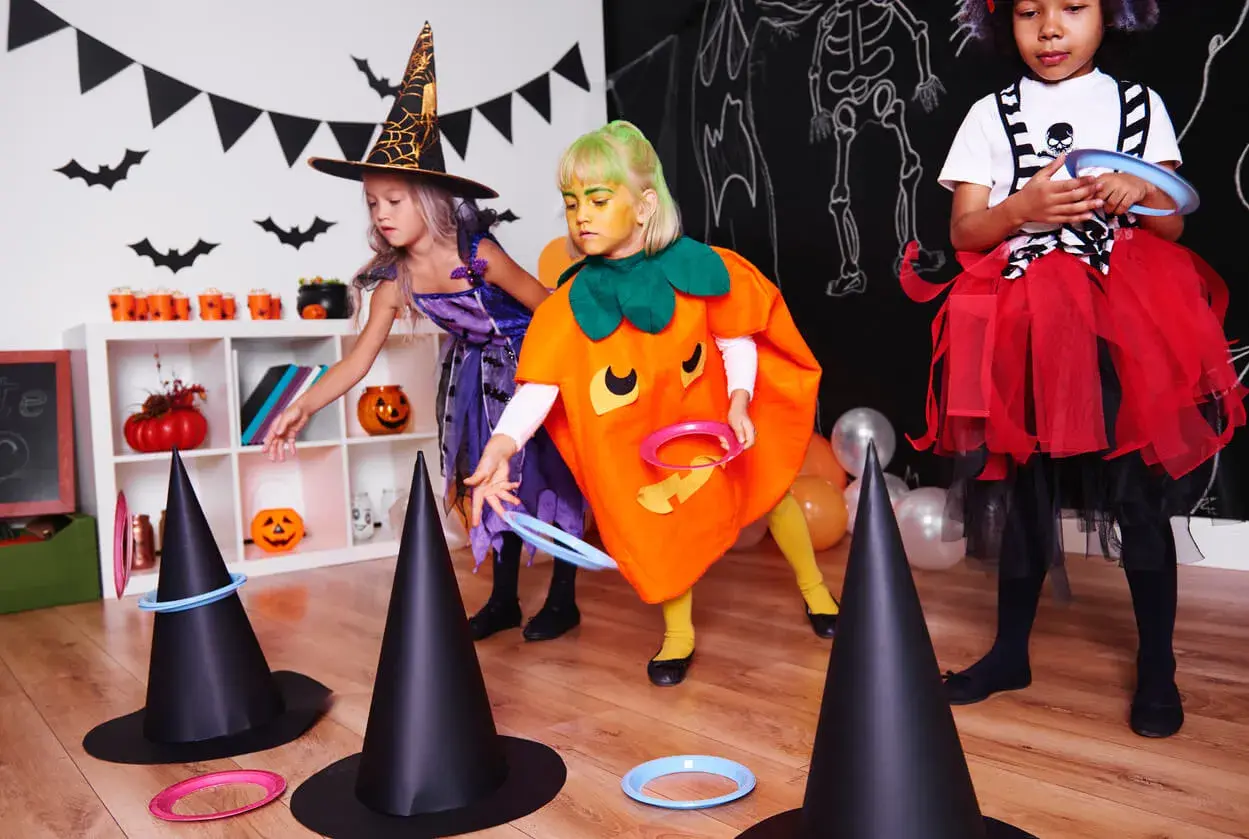 Niñas disfrazadas de bruja y calabaza juegan a lanzar aros en sombreros de bruja, parte de divertidos juegos de Halloween para niños.