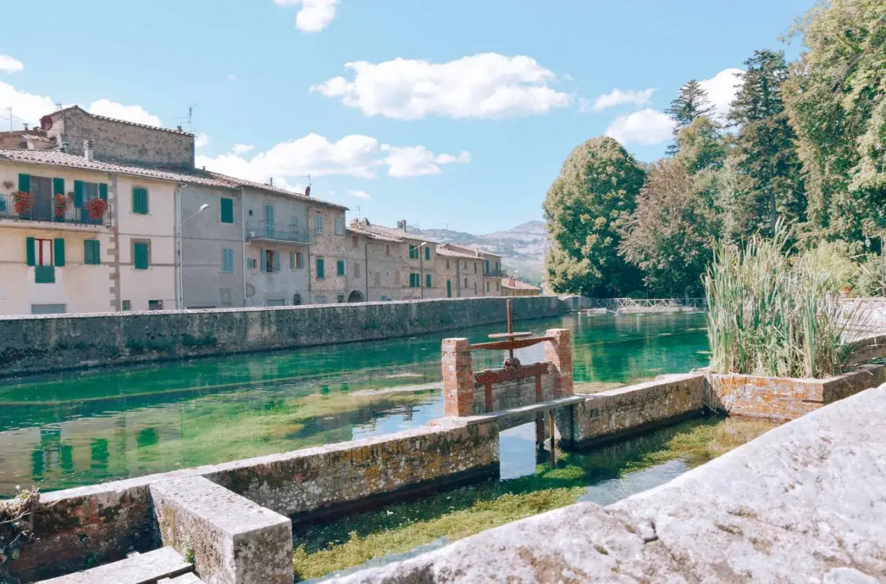 Un canale d'acqua cristallina attraversa un borgo con case antiche, ricordando i pittoreschi **paesi vicini al Monte Amiata**.