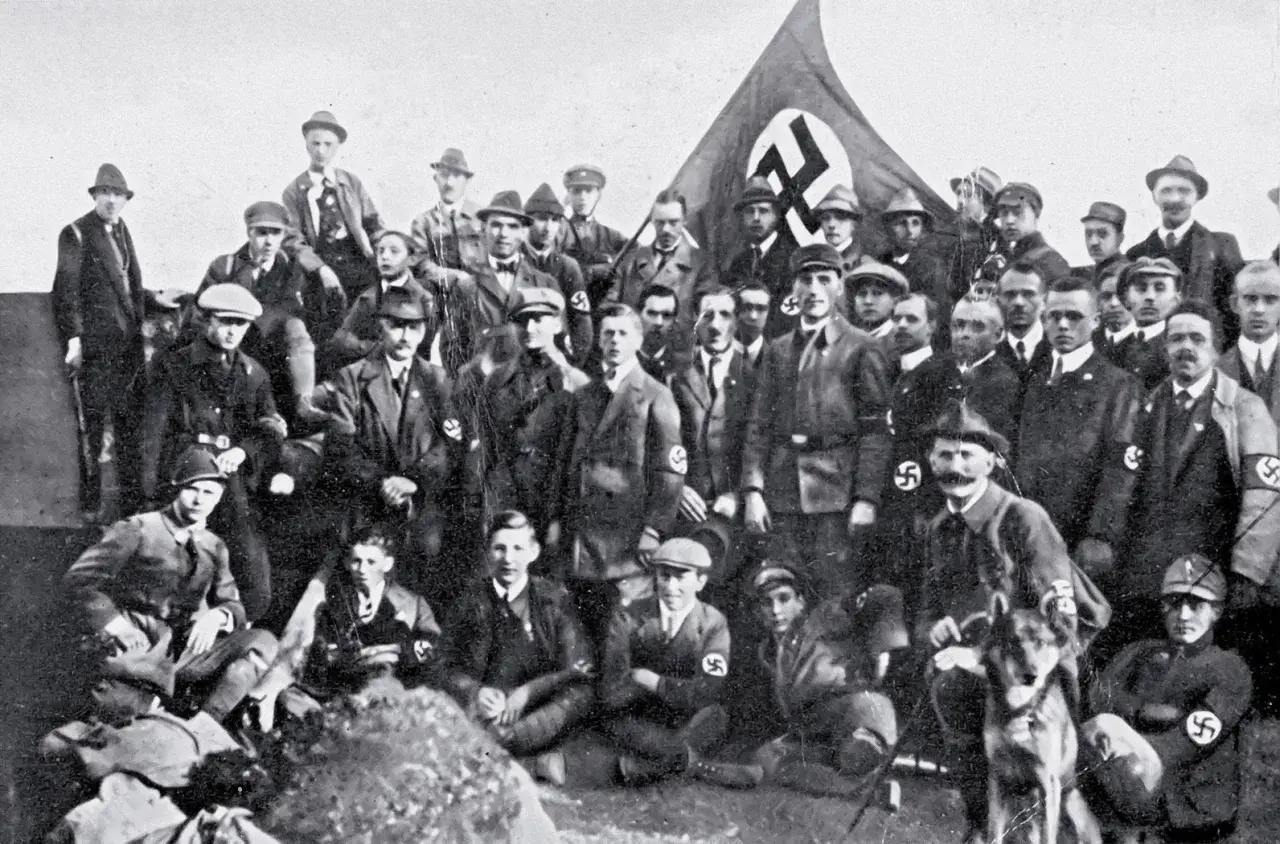 Grupa mężczyzn w mundurach i kapeluszach, z flagą NSDAP w tle. To zdjęcie pokazuje członków partii nazistowskiej, czyli NSDAP.
