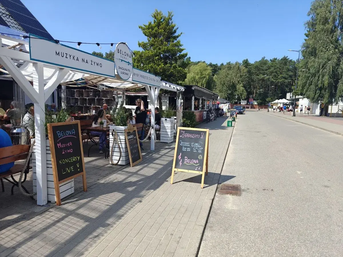 Dębki promenada: lokal Belona zaprasza na muzykę na żywo i koncerty. W tle widać letnie ogródki i spacerujących ludzi.