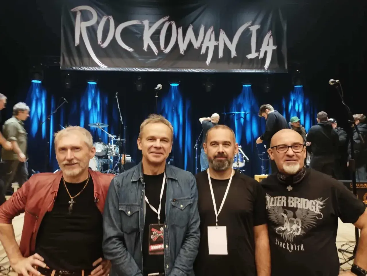 Czterech mężczyzn pozuje na tle sceny z banerem "ROCKOWANIA". Jeden z nich ma na sobie czerwoną kurtkę, inny dżinsową koszulę. W tle widać zespół i sprzęt. Idziemy do przodu dżem!