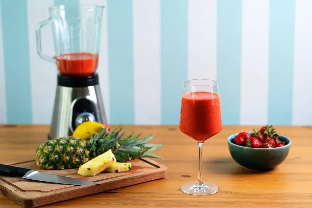 Pyszny koktajl owocowy w kieliszku, obok ananas, truskawki i blender.