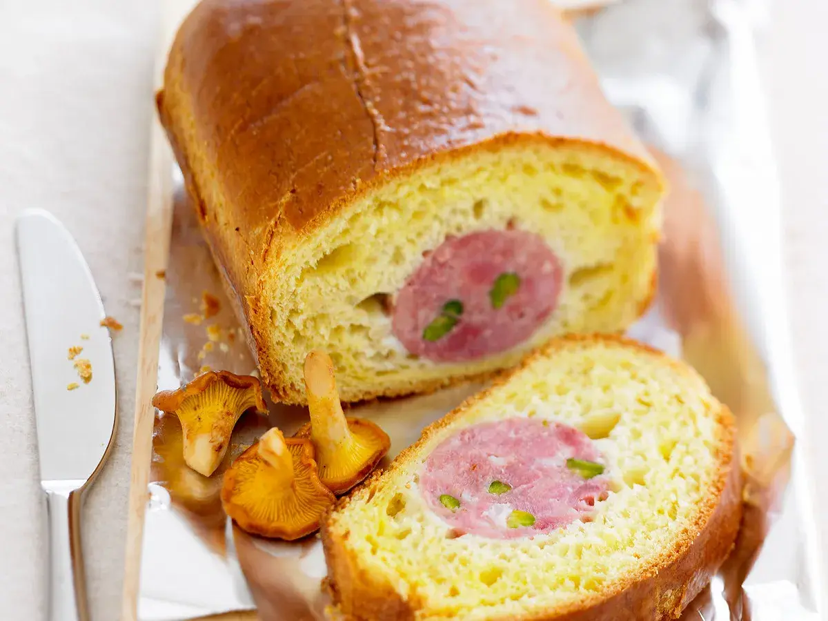 Un saucisson brioché doré, coupé en tranches, révèle sa garniture rosée. Des girolles et un couteau complètent cette gourmandise.