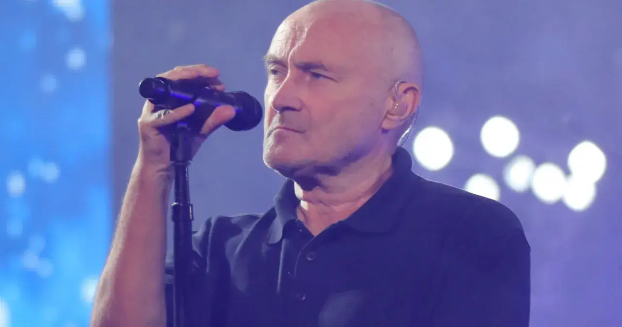 Phil Collins, wokalista zespołu Genesis, śpiewa do mikrofonu na tle niebieskiej sceny z białymi światłami.