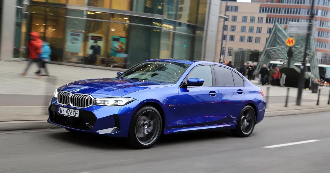 Błękitne BMW 330e mija nowoczesne budynki i wejście do metra.