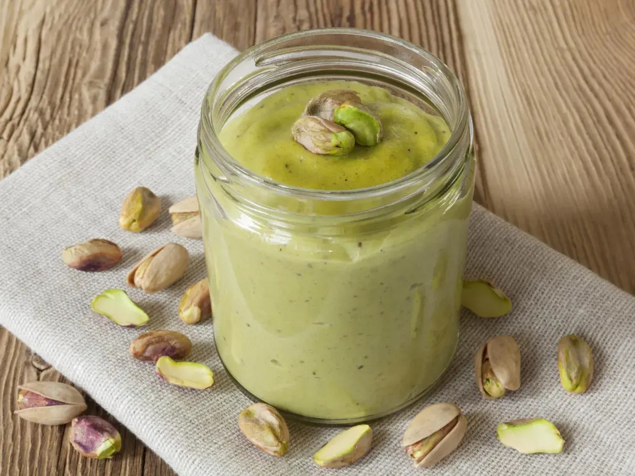 Tarro de crema pistacho casera, decorado con pistachos enteros y trozos, rodeado de más pistachos sobre tela y madera.