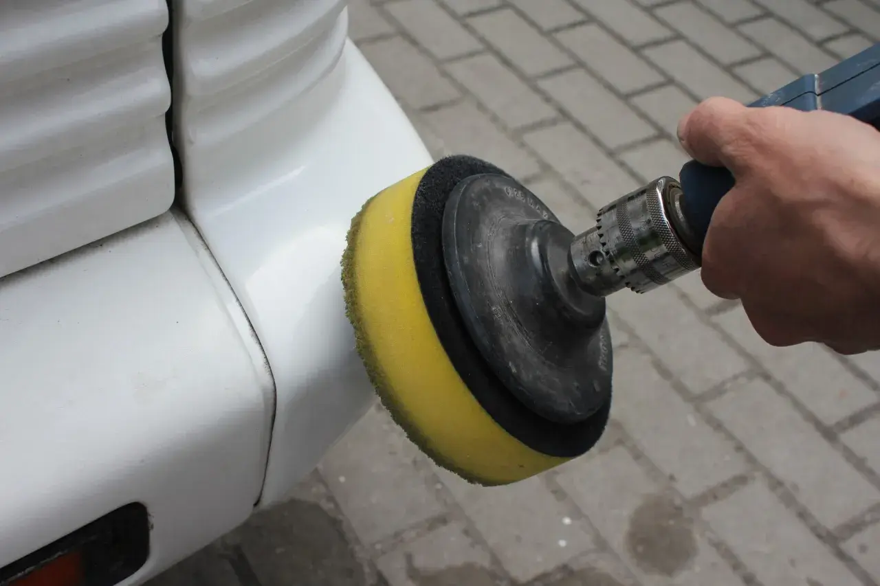 Ręka trzyma polerkę z żółtą gąbką, która jest używana do polerowania białego zderzaka. To pokazuje, jak polerować auto.