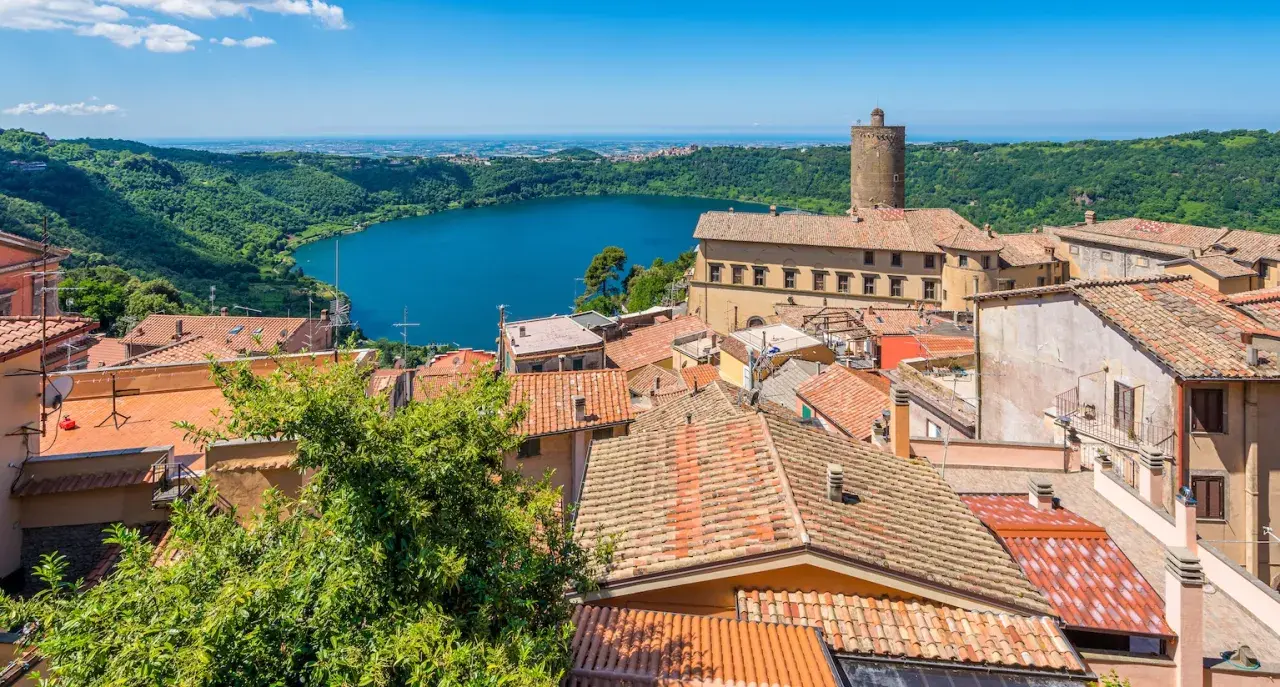Panoramica dei colli di Roma oggi: un lago azzurro circondato da colline verdi e un borgo con tetti rossi e una torre medievale.