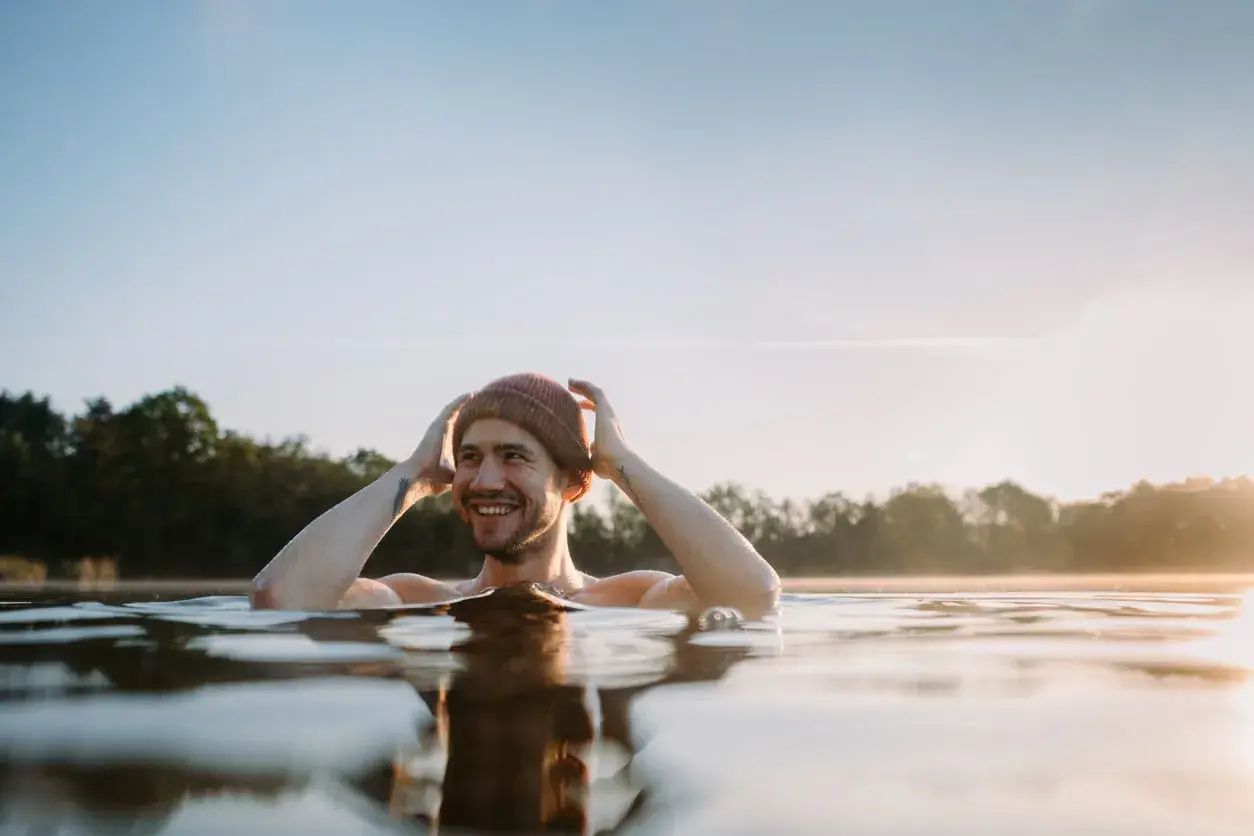 Uśmiechnięty mężczyzna w czapce zanurzony w zimnej wodzie, czerpiący radość z metody wim hof oddychanie.