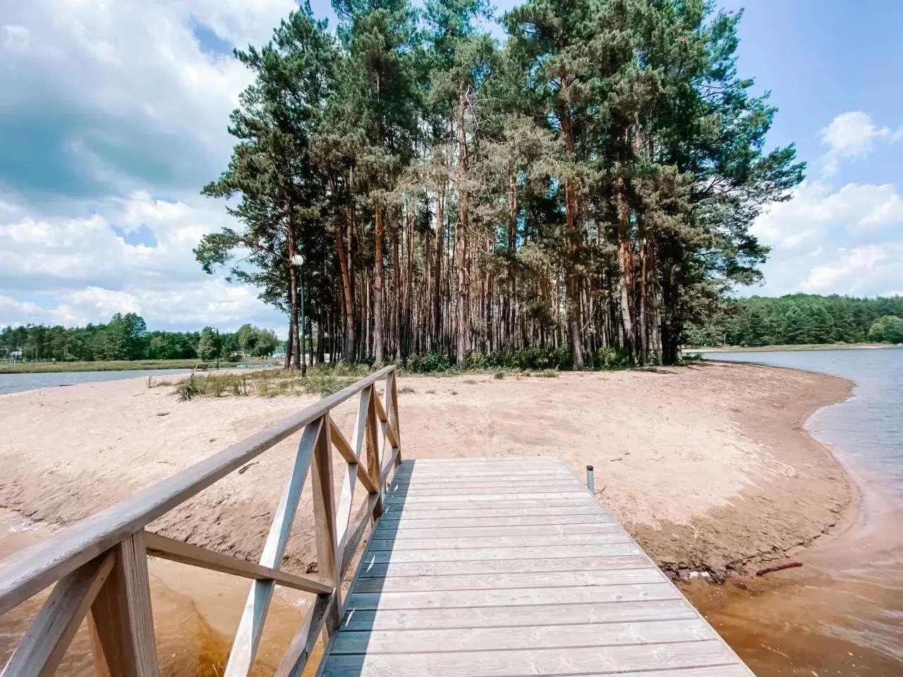 Drewniany pomost prowadzi do piaszczystej plaży otoczonej lasem sosen.
