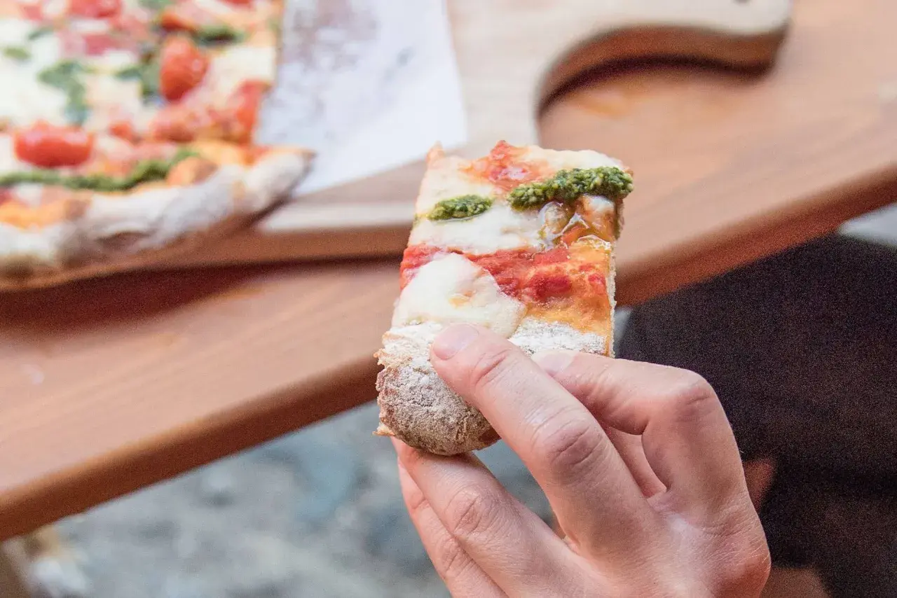 Eine Hand hält ein Stück Pinsa mit Tomaten, Mozzarella und Basilikumpesto. Was ist Pinsa? Eine römische Spezialität, die sich von Pizza unterscheidet.