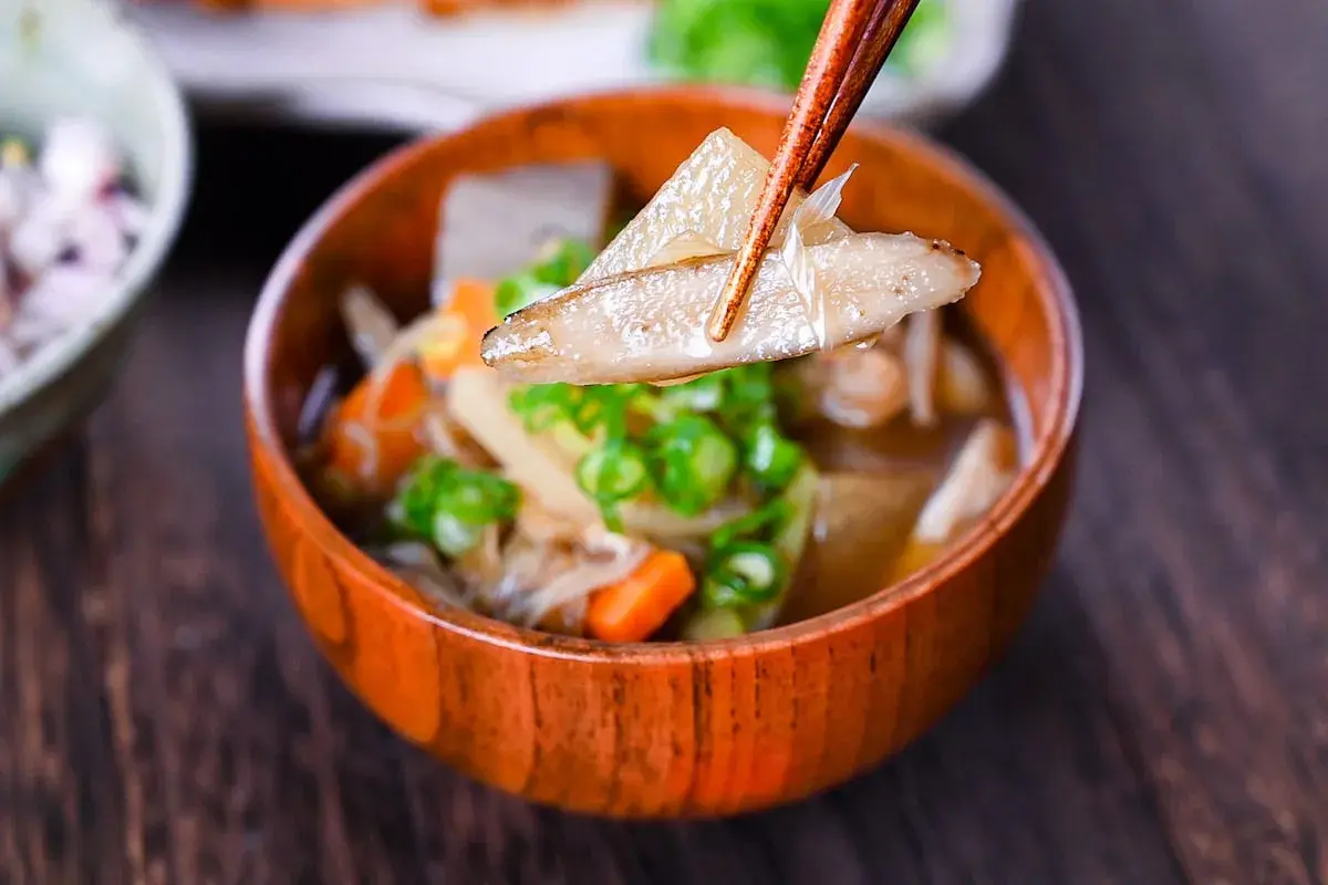 Chopsticks lift a piece of daikon from a steaming bowl of Japanese vegetable soup, filled with carrots, mushrooms, and green onions.