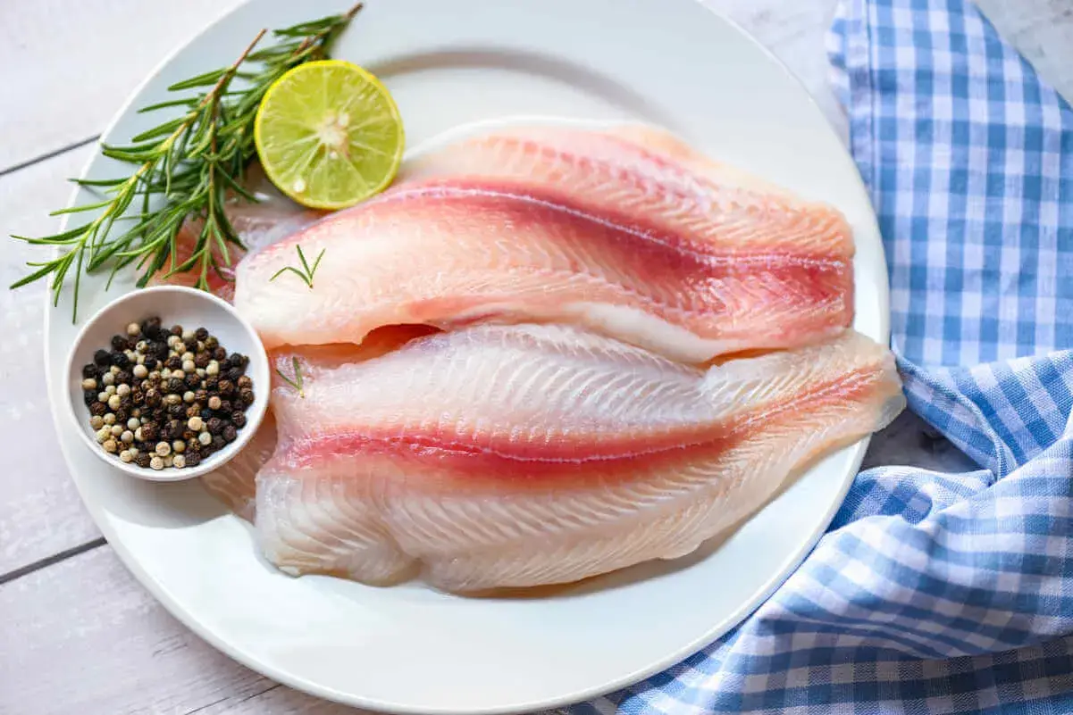 Dos filetes de tilapia frescos, listos para cocinar, acompañados de romero, limón y granos de pimienta.