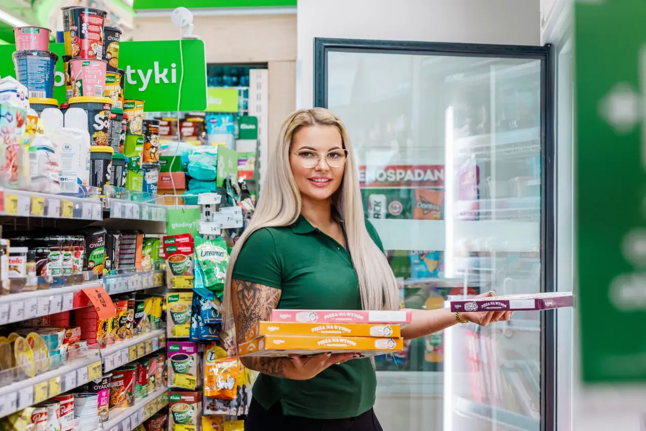 Kobieta w zielonej koszulce trzyma stos pudełek z pizzą na wynos w sklepie osiedlowym.