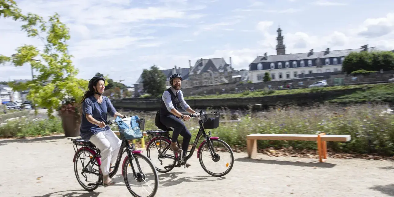 Couple à vélo profitant d'une balade ensoleillée le long de la rivière, avec les bâtiments historiques de Tréguier Lannion en arrière-plan.