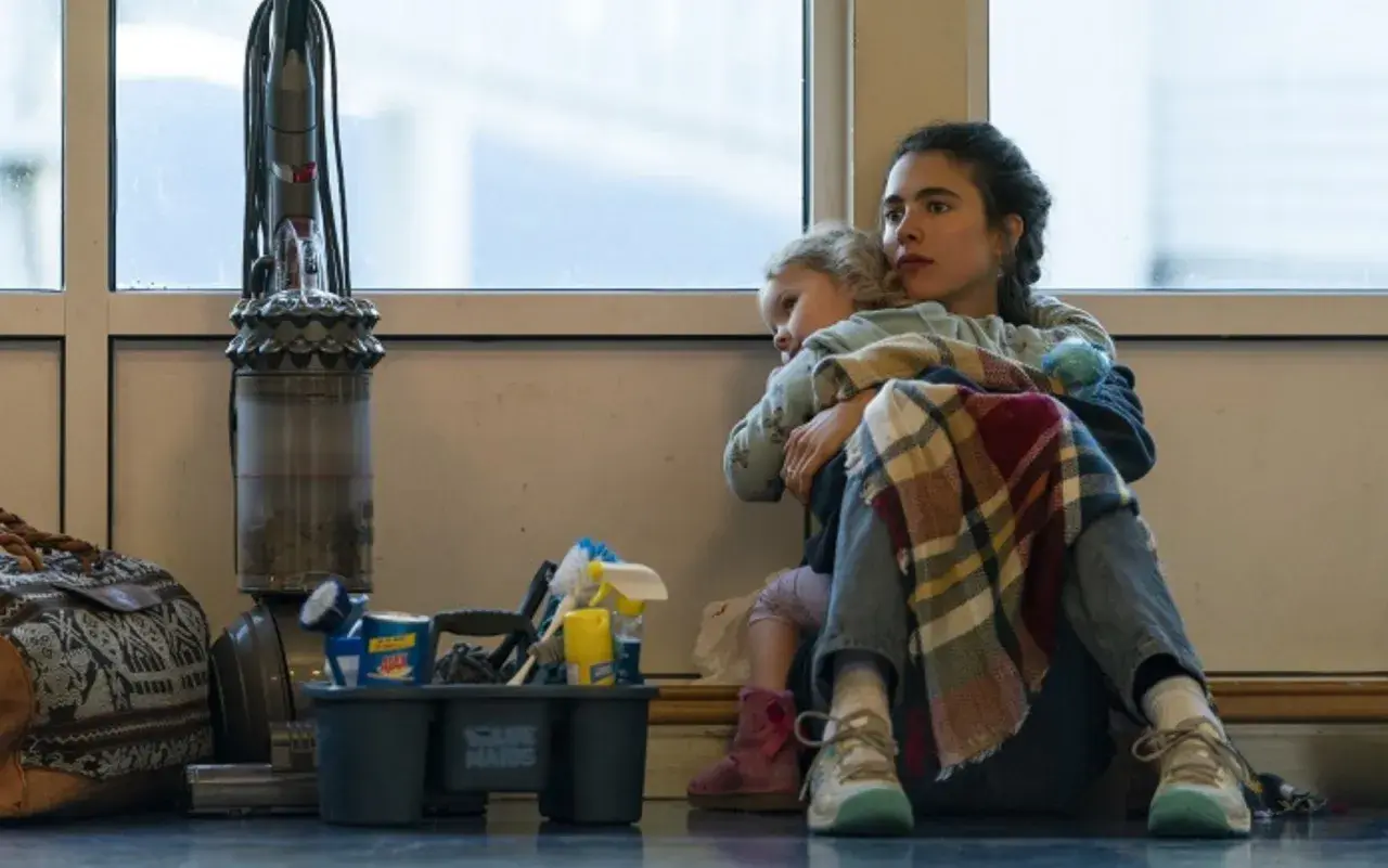 Madre abbraccia figlia, circondata da attrezzi per le pulizie e un aspirapolvere. Le recensioni per il suo lavoro di maid sono positive, ma la vita è dura.