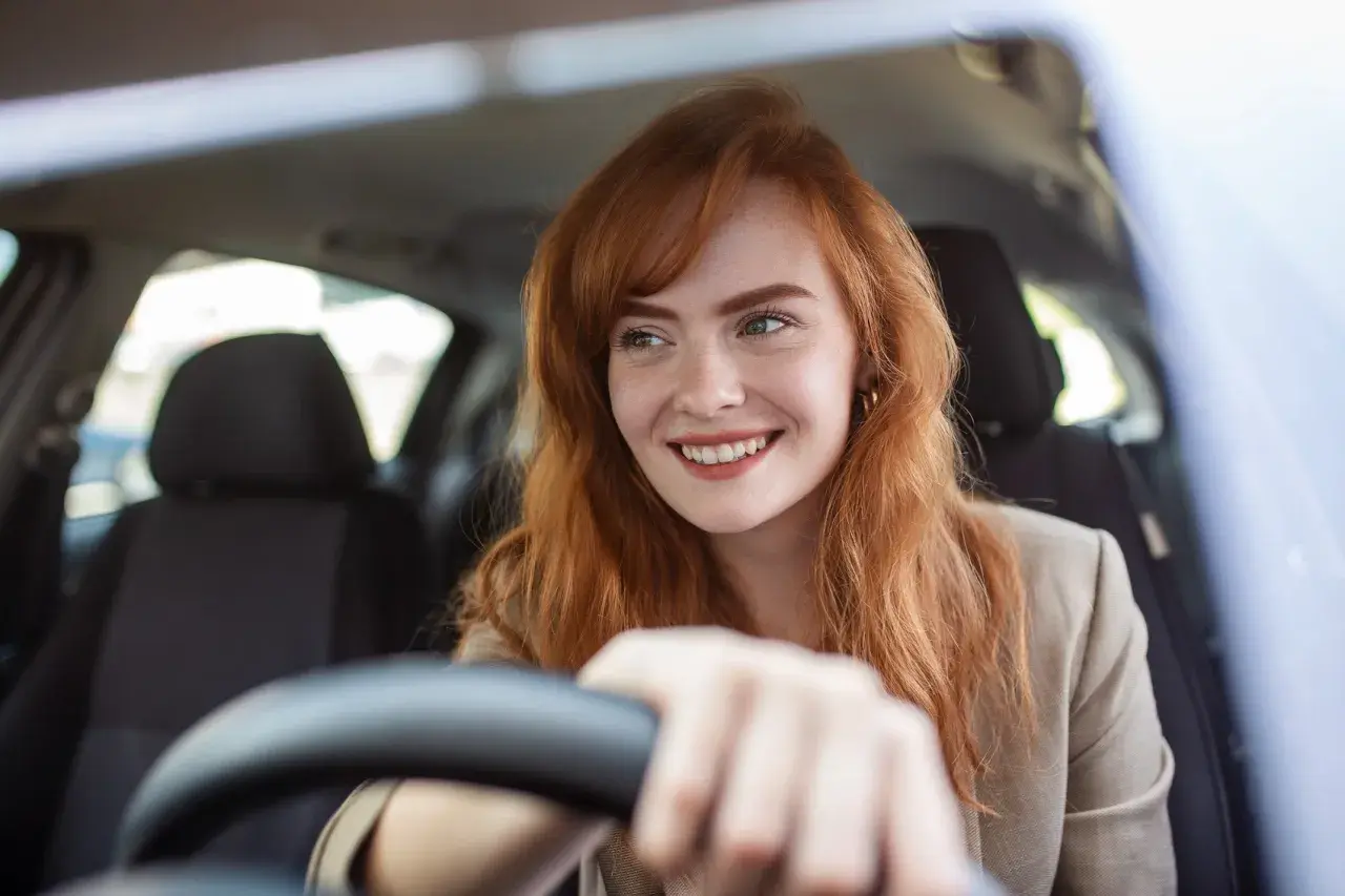 Donna sorridente al volante, con capelli rossi e lentiggini. Pensa alla sua assicurazione giornaliera auto per un viaggio sereno.