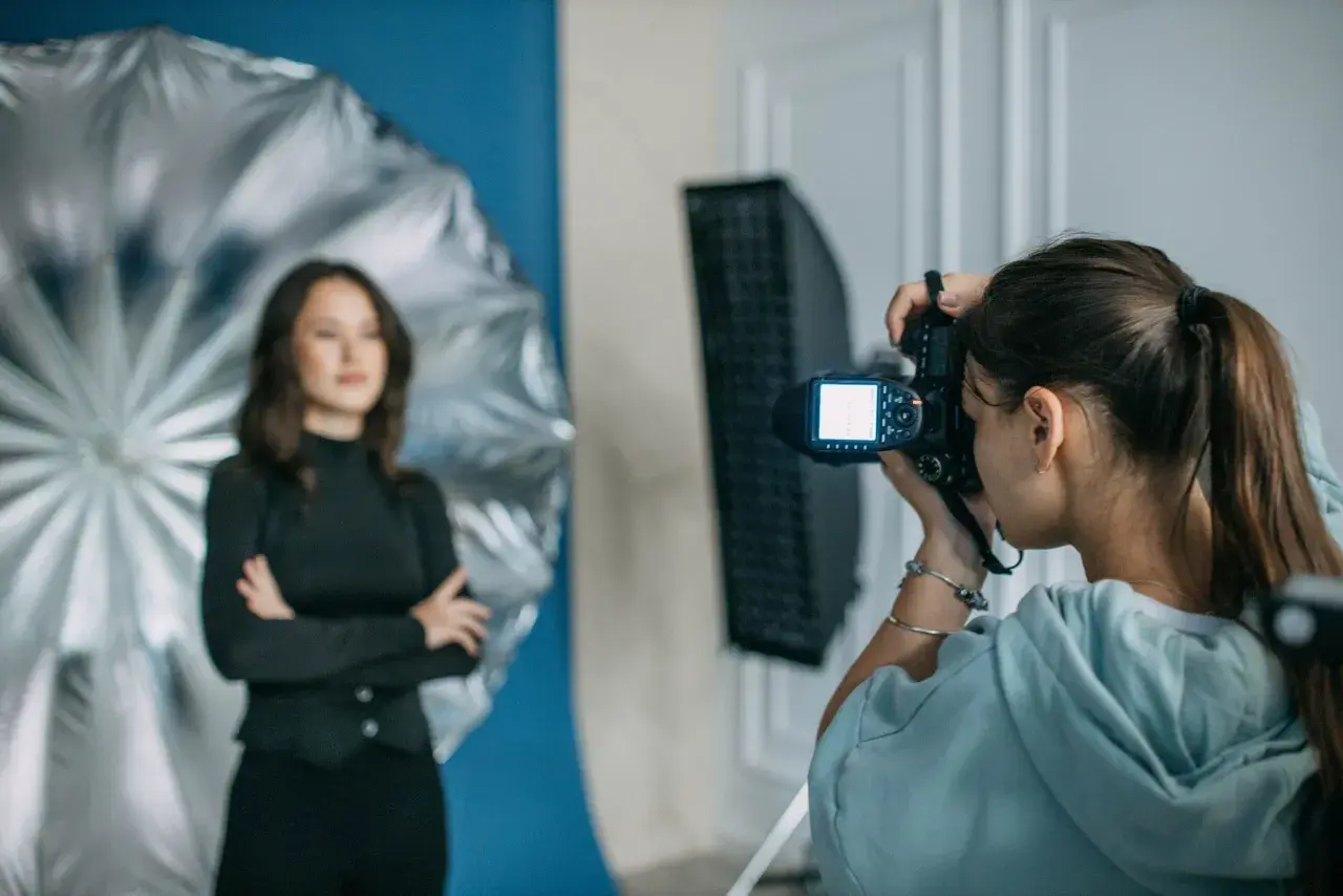 Dzień fotografa w studiu. Młoda kobieta z aparatem w ręku, w tle modelka pozująca na tle niebieskiego tła i srebrnego parasola.