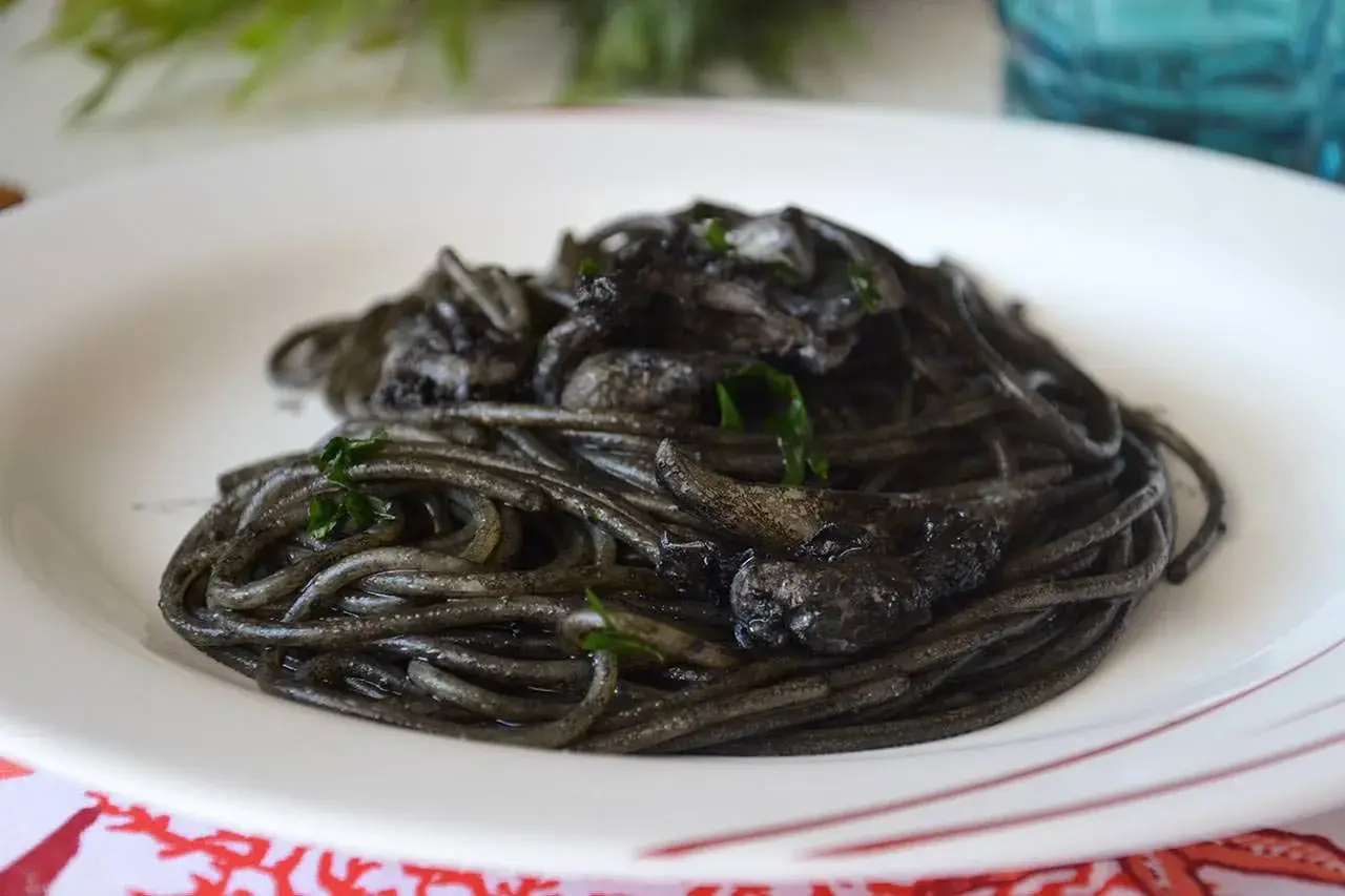 Spaghetti al nero di seppia, un piatto intenso e saporito, servito su un piatto bianco con guarnizioni verdi.