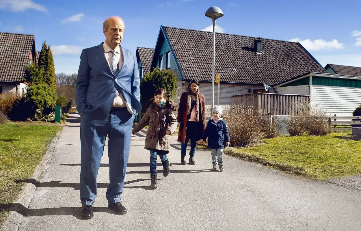 En man i kostym går med en familj i ett villaområde, en scen som kan passa i svenska komedier.