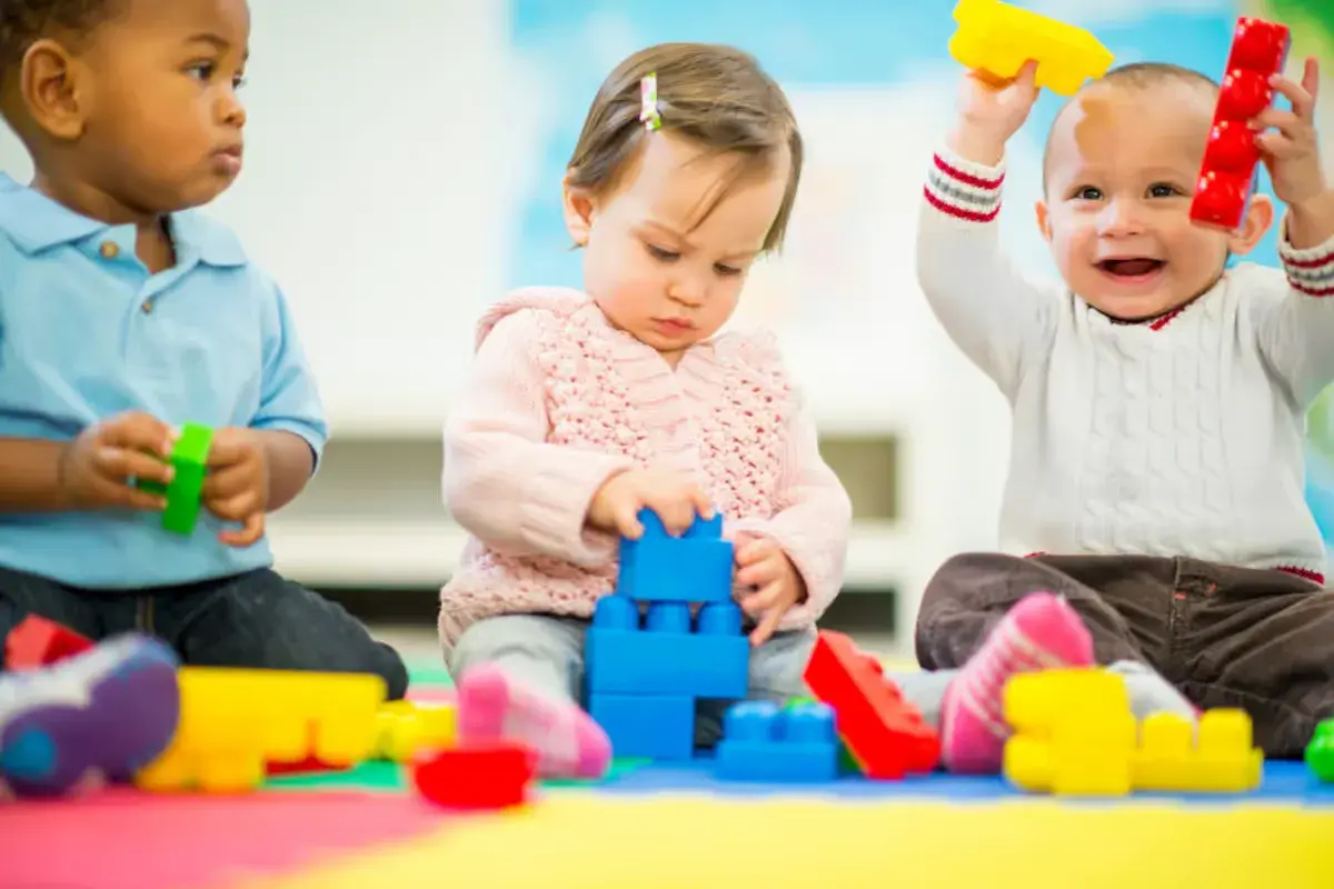 Three toddlers engage in parallel play, each absorbed in their own colorful blocks. One stacks blue blocks, another holds red and yellow toys, and the third plays with green.