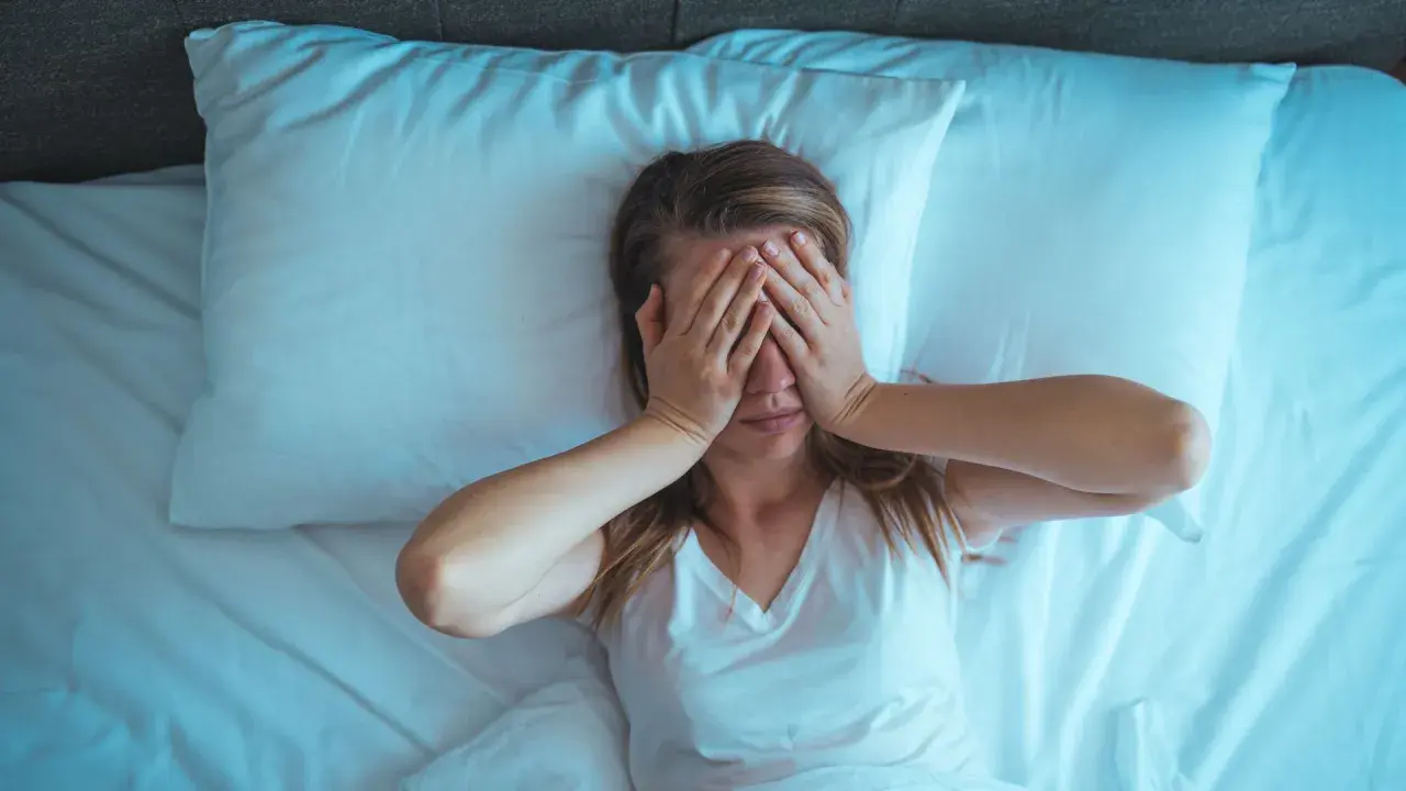 Mujer en la cama, con las manos cubriendo sus ojos. Son las 4 de la mañana y no puedo dormir, la luz azulada ilumina la escena.